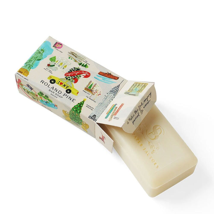 A rectangular off-white Holiday in New York Roland Pine 5 oz Boxed Shea Butter Soap Bar, with embossed text, is partially slid from a colorful illustrated box featuring New York-themed art and hints of cedarwood fragrance.