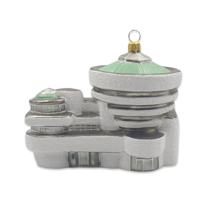 The Guggenheim Building Ornament is a glass decoration inspired by Frank Lloyd Wright’s design, featuring rounded edges, green-tinted domes, and a gold loop at the top for hanging.