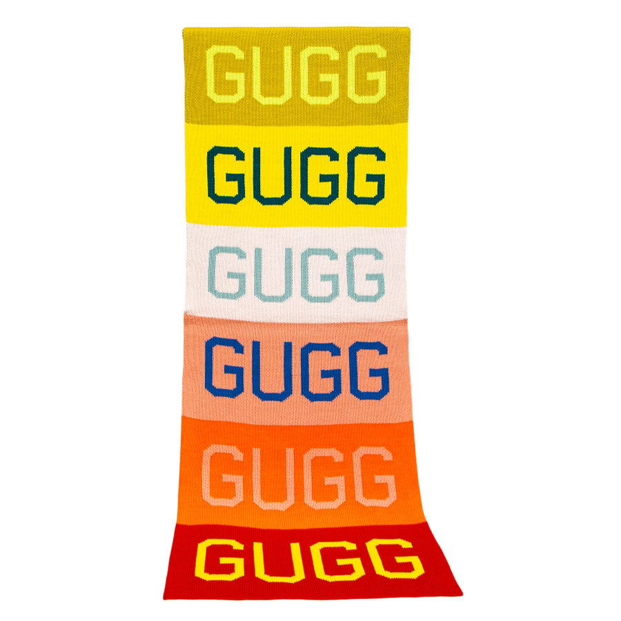 The Gugg Gugg Gugg Scarf is a long, oversized knit with six bold color blocks—yellow, gold, white, blue, orange, and red—each featuring "GUGG" in striking font for a vibrant blend of warmth and style.