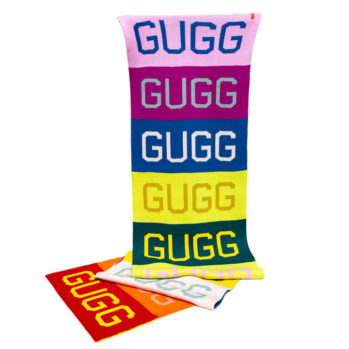 The Gugg Gugg Gugg Scarf features bold block lettering across six vibrant stripes—pink, purple, blue, yellow, green, and yellow—offering colorful warmth and style. Red and light blue folded scarves are displayed underneath.