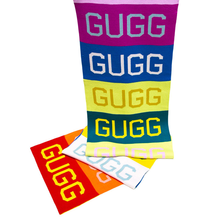 A display of Gugg Gugg Gugg Scarves in bold colors, each featuring "GUGG" in large letters. Stacked and overlapping, these oversized knit scarves combine cozy warmth with standout style.