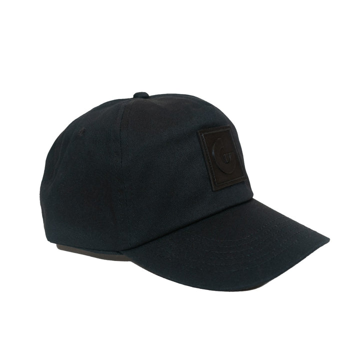 The Guggenheim Constellation Cap New York is a black, adjustable cotton cap with a slightly curved brim and a rectangular embossed logo patch on the front, showcasing its minimalist style against a plain white background.