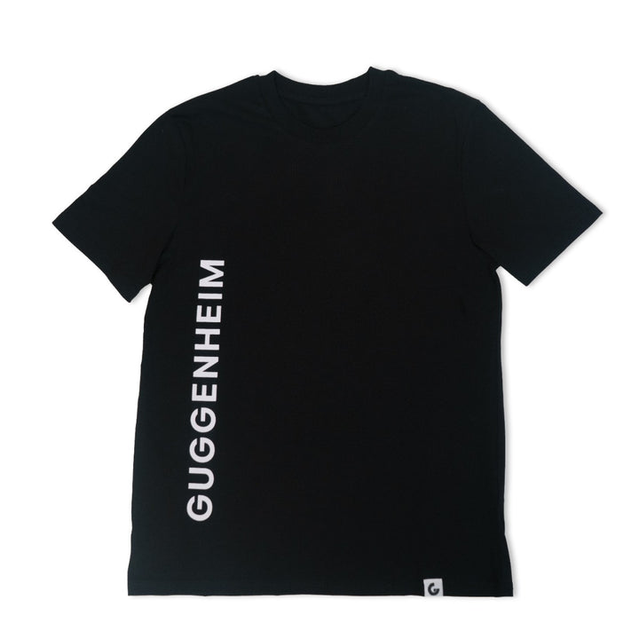The Guggenheim Constellation T-Shirt, New York Black, features short sleeves, organic cotton fabric, and “GUGGENHEIM” printed vertically in white on the left side with a G logo tag at the hem—ideal for fans of iconic art institutions.