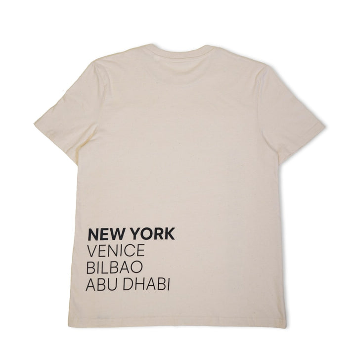 The Guggenheim Constellation T-Shirt, New York Natural, is an organic cotton tee in off-white. Laid flat, its lower left back features black all-caps city names—NEW YORK, VENICE, BILBAO, ABU DHABI—inspired by contemporary art.