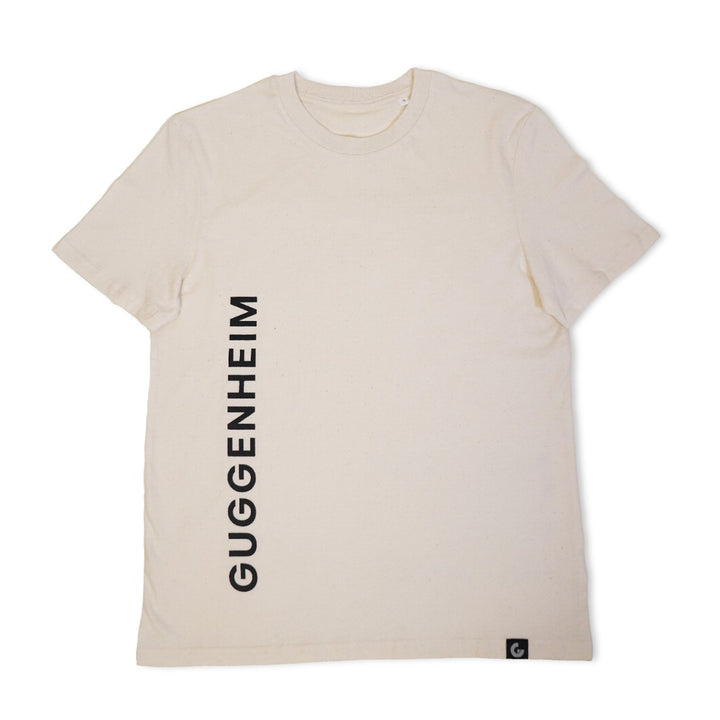The Guggenheim Constellation T-Shirt, New York Natural, is an off-white organic cotton tee featuring "GUGGENHEIM" printed vertically in black on the left side and a small black tag at the lower hem—ideal for art lovers.
