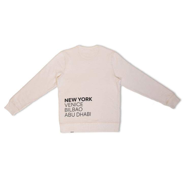 The Guggenheim Constellation Crewneck Sweatshirt, New York Natural, is a light organic cotton sweatshirt featuring NEW YORK, VENICE, BILBAO, ABU DHABI in black on the lower left front, shown laid flat on a white background.