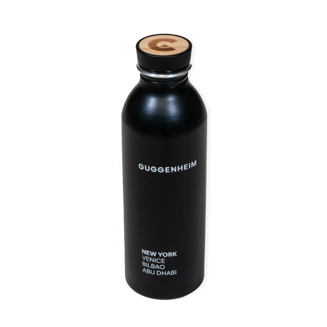 The Guggenheim Constellation Water Bottle New York is a stylish black recycled aluminum bottle, with "Guggenheim" in white and New York, Venice, Bilbao, Abu Dhabi below. The cap features a wooden top with the G logo.