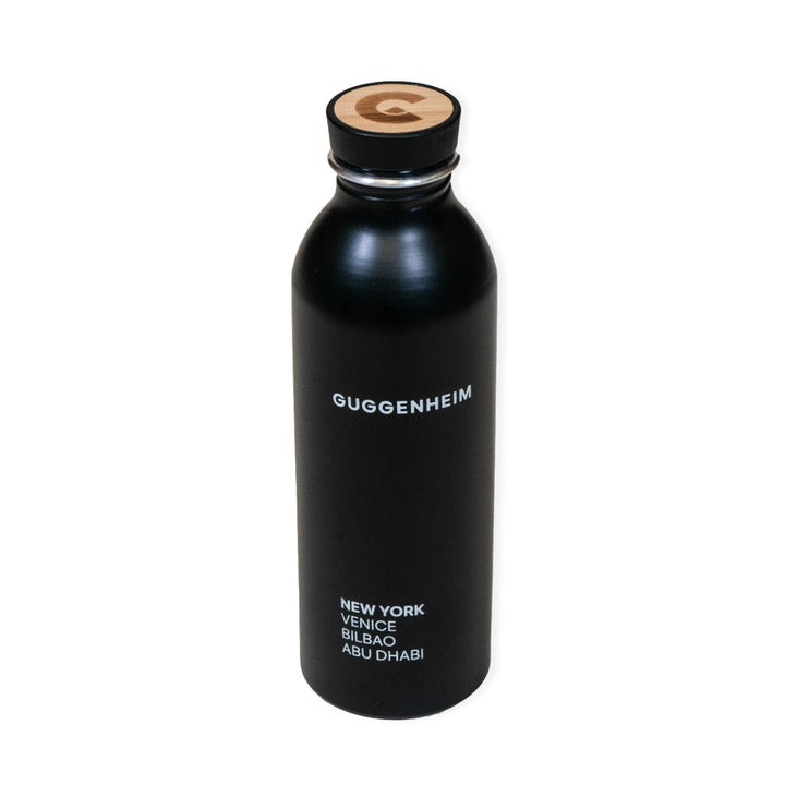 The Guggenheim Constellation Water Bottle New York is a stylish black recycled aluminum bottle, with "Guggenheim" in white and New York, Venice, Bilbao, Abu Dhabi below. The cap features a wooden top with the G logo.