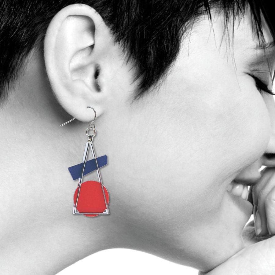 A person with short dark hair smiles with eyes closed, hand near face. They wear Kandinsky Earrings, Triangle At Rest, featuring a red circle, blue rectangle, and silver triangle. The image is mostly black and white.