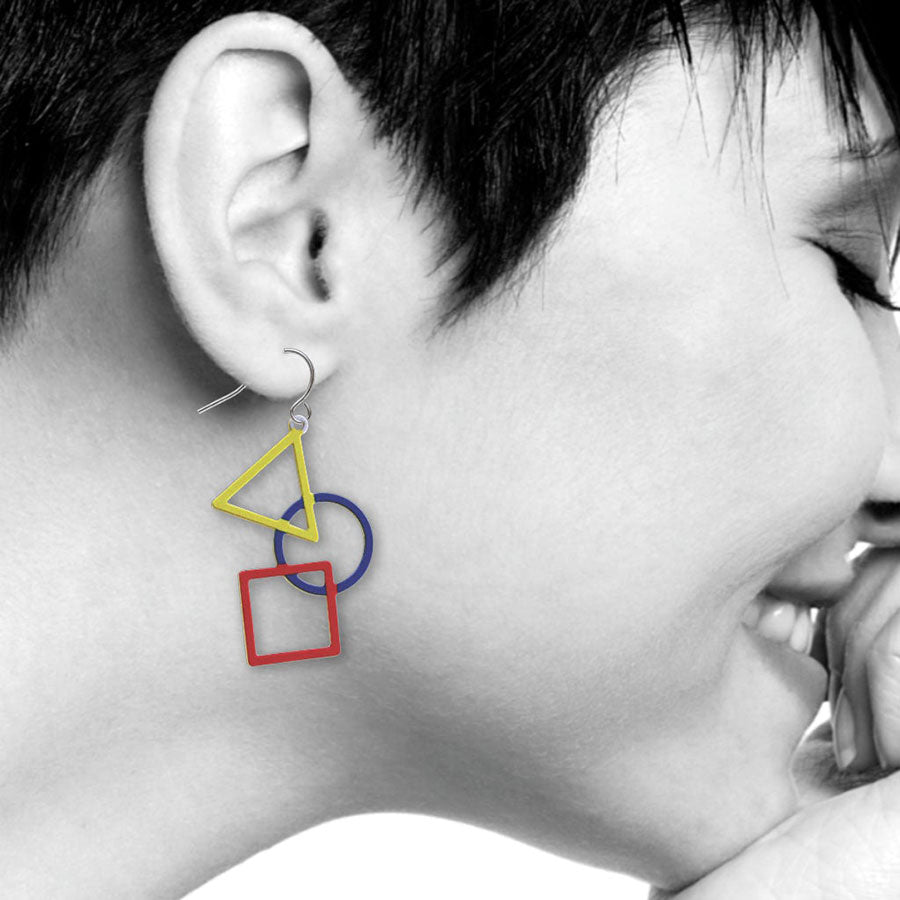 A close-up of a smiling person with eyes closed, wearing Kandinsky Earrings—Triangle, Circle & Square—showcasing vibrant primary colors against a mostly black-and-white background.