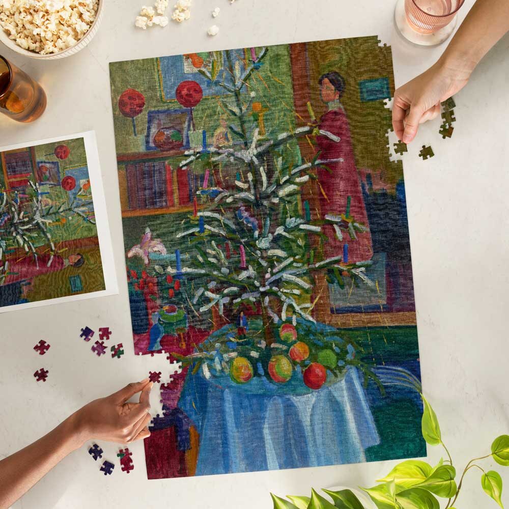 Two people assemble the Gabriele Münter, Christmas Still Life Puzzle—a colorful 1000-piece puzzle featuring a decorated tree and fruit in a cozy room. The puzzle box and matching reference poster are nearby on the white table.
