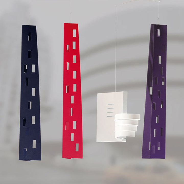 Four hanging paper models echo the New York City skyline: three are tall and narrow (blue, red, purple), while one is a boxy white shape reminiscent of the Guggenheim Building Mobile by Flensted.