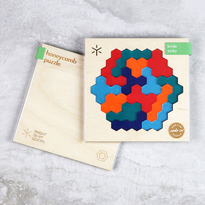 Two square puzzles are shown: a colorful honeycomb puzzle made of hexagonal pieces, labeled “kinda tricky,” and the packaging for the Adventure Honeycomb Puzzle. Both rest on a gray surface, perfect for your coffeetable.