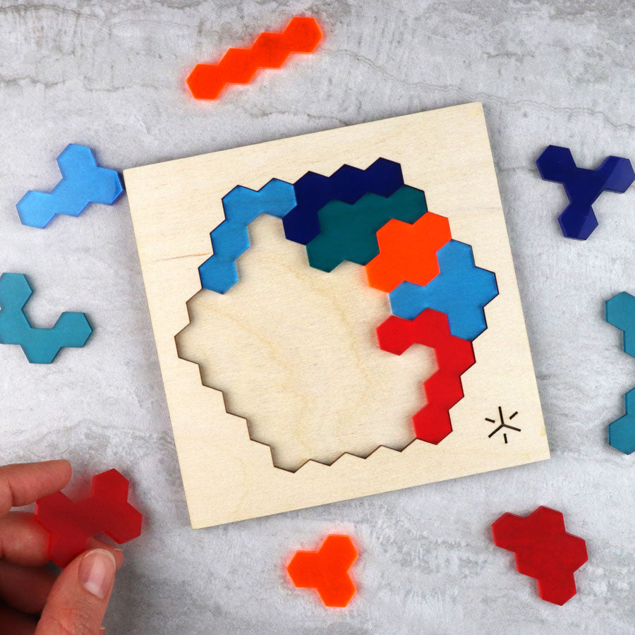 A hand places a red piece into the wooden Adventure Honeycomb Puzzle, which features multicolored hexagonal pieces. Several unused pieces from the puzzle are scattered nearby on a gray surface.