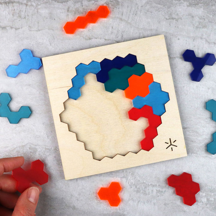 A hand places a red piece into the wooden Adventure Honeycomb Puzzle, which features multicolored hexagonal pieces. Several unused pieces from the puzzle are scattered nearby on a gray surface.