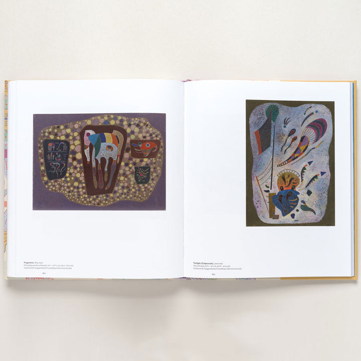 An open book from "Vasily Kandinsky: Around the Circle" shows two abstract, colorful paintings inspired by European modernism on white-bordered pages with captions—organic shapes on purple left, swirling forms and lines right.