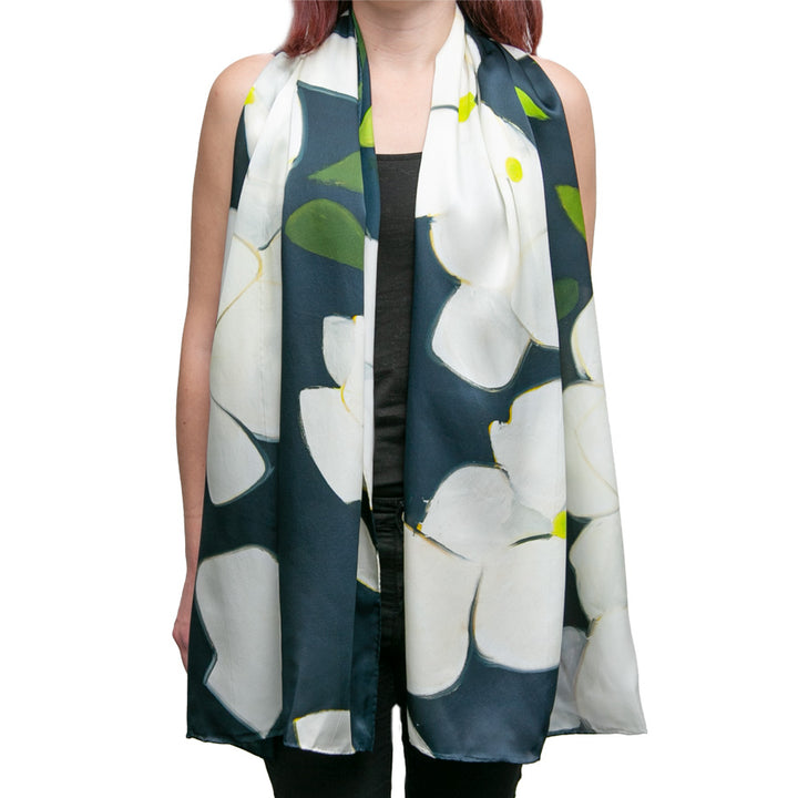A woman wears the Alex Katz Dogwood Silk Scarf, featuring a bold white flower and green-yellow leaves on a dark background, draped around her neck over a black top. Her face is not visible.
