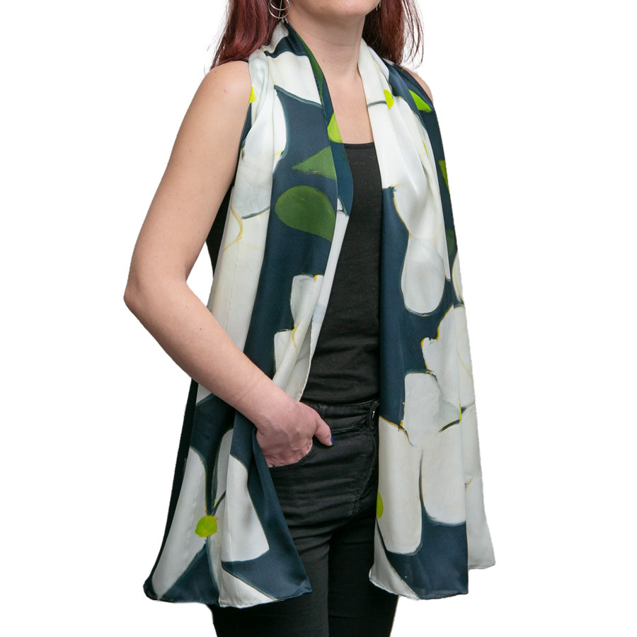 A woman wears a sleeveless black top and pants with a long, flowy Alex Katz Dogwood Silk Scarf featuring a bold white and green floral print draped around her neck.