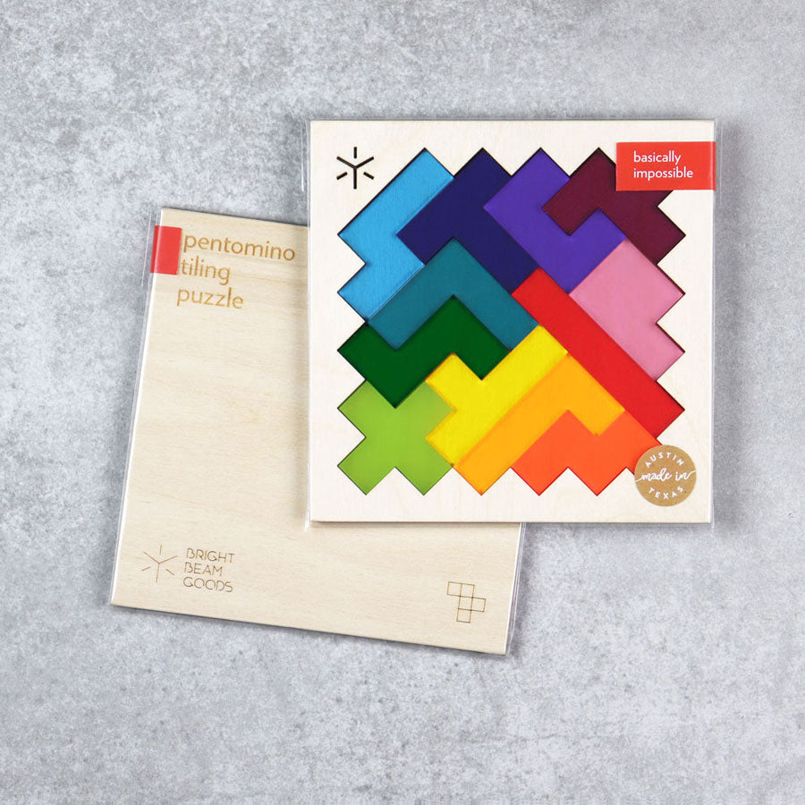 The Rainbow Pentomino Puzzle features two wooden boards: one with vibrant interlocking pieces labeled "basically impossible," offering a real coffee table challenge, and one displaying the puzzle’s plain wooden back.