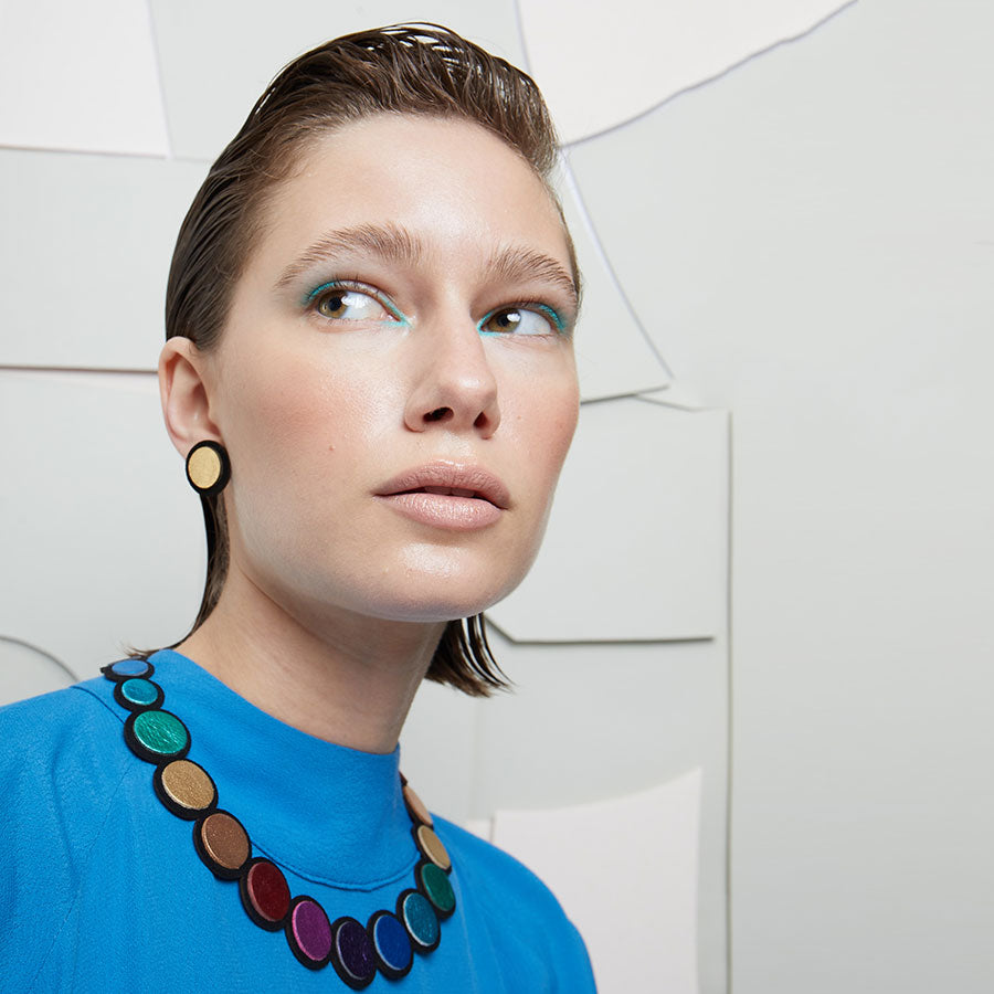 A woman with slicked-back hair poses against a white geometric background, wearing a bright blue top, light blue eye makeup, and the Rainbow Circles Necklace by Iskin Sisters with matching earrings.