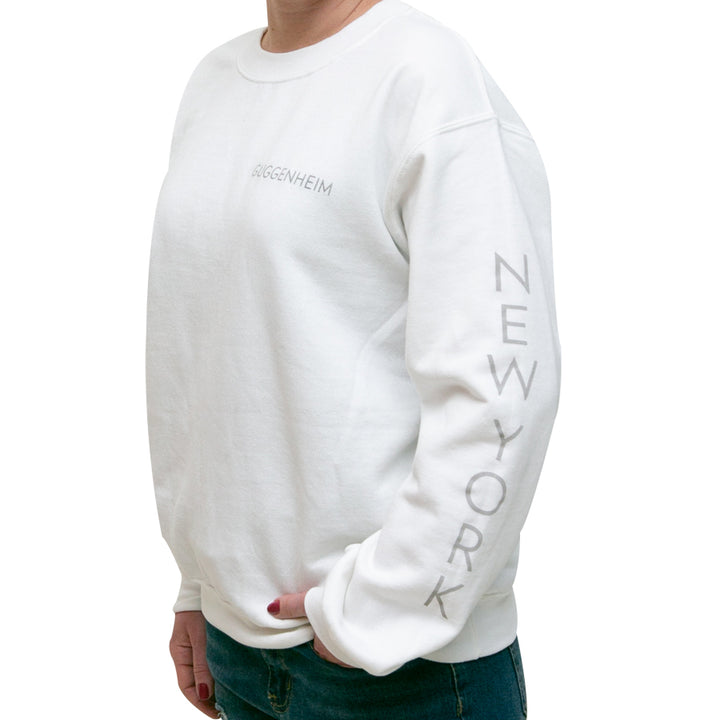 A person stands sideways with one hand in their pocket, wearing the unisex Guggenheim Rotunda Sweatshirt featuring a cotton crew neck fleece, "GUGGENHEIM" embroidered on the chest, and "NEW YORK" printed down the left sleeve.
